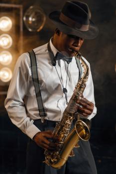 Black jazzman in hat plays the saxophone on the stage with spotlights. Black jazz musician preforming on the scene