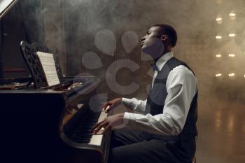Ebony grand piano player playing on the stage with spotlights on background. Negro performer poses at musical instrument before concert