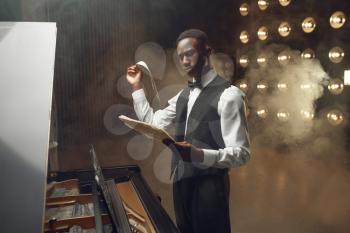 Ebony grand piano player with music notebook in his hands on the stage with spotlights on background. Negro performer poses at musical instrument before concert