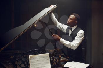 Ebony player tuning grand piano on the stage with spotlights on background. Negro performer poses at musical instrument before concert