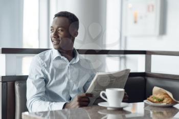 Young ebony businessman with newspaper having lunch in office cafe. Successful business person drinks coffee in food-court, black man in formal wear