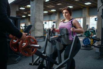 Overweight woman on exercise machine in gym, training with instructor. Female person struggles with excess weight, aerobic workout against obesity
