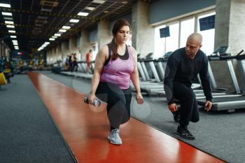 Overweight woman holds dumbbells, exercise with trainer in sport club, fit training with instructor. Female person struggles with excess weight, aerobic workout against obesity, gym