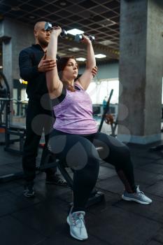 Overweight woman with trainer doing exercise with dumbbells in sport club, fitness training with instructor. Female person struggles with excess weight, aerobic workout against obesity, gym