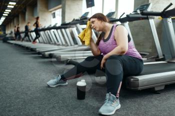 Tired overweight woman sitting on treadmill in gym, leisure after active training. Obese female person struggles with excess weight, aerobic workout against obesity, sport club