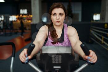 Overweight woman trains on exercise bike in gym, active training. Obese female person struggles with excess weight, aerobic workout against obesity, sport club