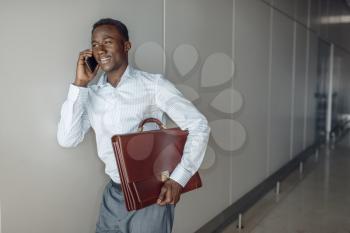 Ebony businessman with briefcase talking by phone in office hallway. Successful business person negotiates in corridor, black man in formal wear