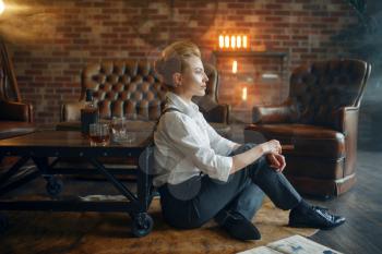Woman in strict clothes sitting on the floor with whiskey and cigar, retro fashion, gangster style. Vintage business lady in office with brick walls