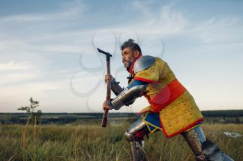 Old medieval knight in armor holds axe, great tournament. Armored ancient warrior in armour posing in the field