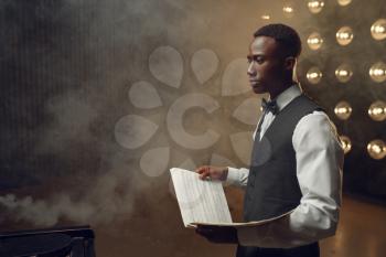 Black pianist with music notebook in his hands on the stage with spotlights on background. Black performer poses at musical instrument before concert