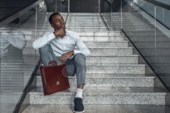 Black businessman with briefcase sitting on stairs, mall. Successful business person, black man in formal wear, shopping center