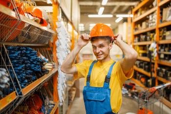 Male builder trying on helmet at the shelf in hardware store. Constructor in uniform look at the goods in diy shop
