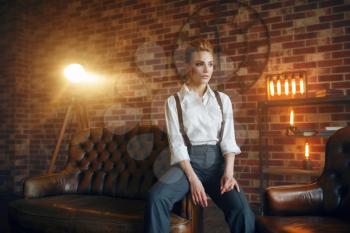 Sexy business woman in strict clothes poses on leather couch in studio, retro fashion, gangster style, mafia. Vintage lady in office with brick walls