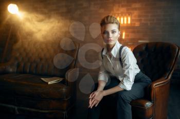 Elegance business woman in strict clothes poses on leather couch in studio, retro fashion, gangster style, mafia. Vintage lady in office with brick walls