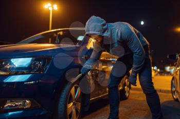 Car robber punctures the tyre, vandalism, hooligan. Hooded male bandit spoils vehicle on parking. Auto robbery, automobile crime