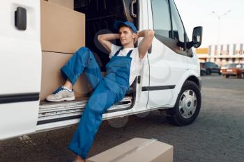 Deliveryman in uniform relaxing in the car during a break, auto with parcels and carton boxes, delivery service. Man poses at cardboard packages in vehicle, male deliver, courier or shipping job