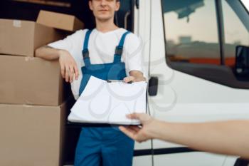 Deliveryman in uniform holding parcel and notebook, carton boxes in the car, delivery service. Man standing at cardboard packages in vehicle, male deliver