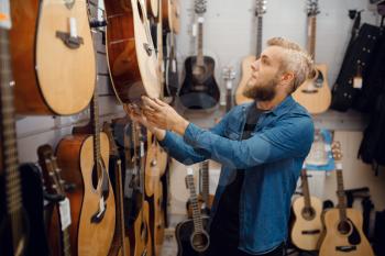 Bearded young man choosing acoustic guitar in music store. Assortment in musical instruments shop, male musician