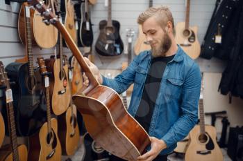 Bearded young man choosing acoustic guitar in music store. Assortment in musical instruments shop, male musician buying equipment