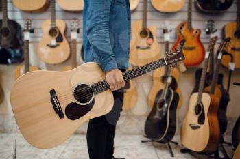 Young man holding acoustic guitar in music store. Assortment in musical instruments shop, male musician buying equipment