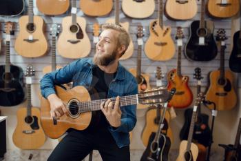 Young man plays on acoustic guitar in music store. Assortment in musical instruments shop, male musician buying equipment