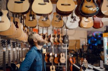 Bearded young guy choosing acoustic guitar in music store. Assortment in musical instruments shop, male musician buying equipment