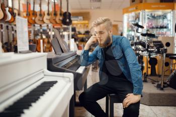 Young male musician poses at the piano in music store. Assortment in musical instruments shop, keyboardist buying equipment, pianist in market