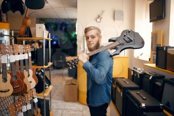 Young man poses with electric guitar on his shoulder in music store. Assortment in musical instruments shop, male musician buying equipment