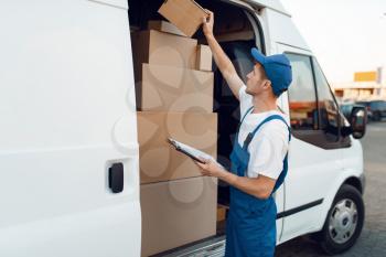 Deliveryman in uniform check boxes in the car, delivery service. Man standing at cardboard packages in vehicle, male deliver