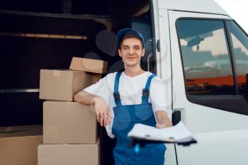 Deliveryman in uniform holding parcel and notebook, carton boxes in the car, delivery service. Man standing at cardboard packages in vehicle, male deliver