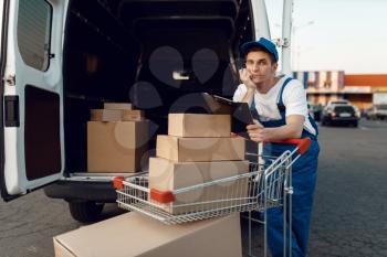 Deliveryman in uniform holds cart with boxes at the car with parcels, delivery service. Man standing at cardboard packages in vehicle, male deliver, courier or shipping job