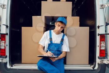 Deliveryman in uniform, parcels in the car on background, cargo, delivery service. Man standing at cardboard packages in vehicle, male deliver, courier or shipping job