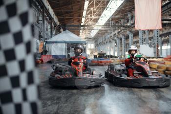 Two kart racers on start line, checkered flag, front view, karting auto sport indoor. Speed race on close go-cart track with tire barrier. Fast vehicle competition, high adrenaline leisure