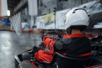 Kart racer in helmet, back view, karting auto sport indoor. Speed race on close go-cart track with tire barrier. Fast vehicle competition, high adrenaline leisure