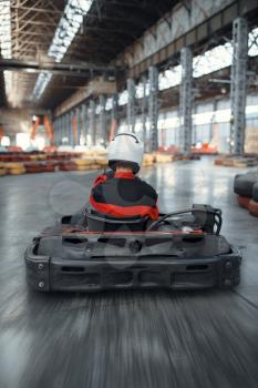 Racer in helmet driving go kart car, back view, karting auto sport indoor. Speed race on close go-kart track with tire barrier. Fast vehicle competition, hot pursuit