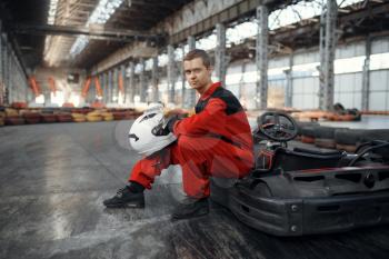 Kart racer in red uniform poses with hemet in hands, karting auto sport indoor. Speed race on close go-kart track with tire barrier. Fast vehicle competition, high adrenaline hobby