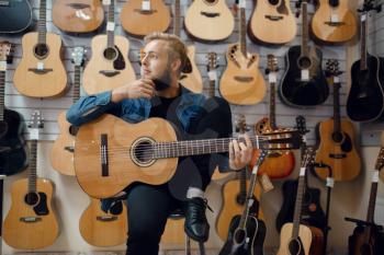 Young man plays on acoustic guitar in music store. Assortment in musical instruments shop, male musician buying equipment