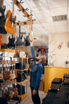 Bearded young man choosing acoustic guitar in music store. Assortment in musical instruments shop, male musician buying equipment