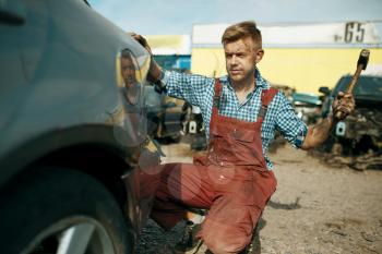 Male repairman works with hammer on car junkyard. Auto scrap, vehicle junk, automobile garbage, abandoned, damaged and crushed transport