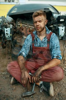 Male repairman sitting on the ground on car junkyard. Auto scrap, vehicle junk, automobile garbage, abandoned, damaged and crushed transport