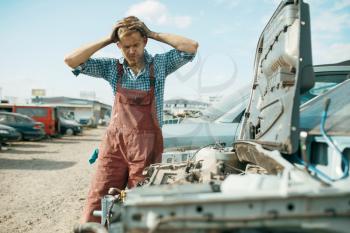 Angry male repairman works on car junkyard. Auto scrap, vehicle junk, automobile garbage, abandoned, damaged and crushed transport