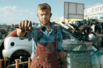 Male repairman shows wrench on car scrapyard. Auto scrap, vehicle junk, automobile garbage. Abandoned, damaged and crushed transport, junkyard