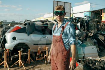 Male repairman in welding glasses on car scrapyard. Auto scrap, vehicle junk, automobile garbage. Abandoned, damaged and crushed transport, junkyard