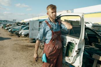 Male repairman holds door on car junkyard. Auto scrap, vehicle junk, automobile garbage. Abandoned, damaged and crushed transport, scrapyard