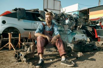Male repairman sitting on the ground on car junkyard. Auto scrap, vehicle junk, damaged and crushed transport