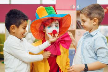Little joyful boy touches red clown's nose, children playing together. Birthday party in playroom, baby holiday in playground. Childhood happiness, childish leisure