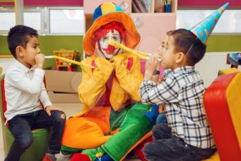 Funny clown with cheerful children blows into the tubes together. Birthday party celebrating in playroom, baby holiday in playground. Childhood happiness, childish leisure