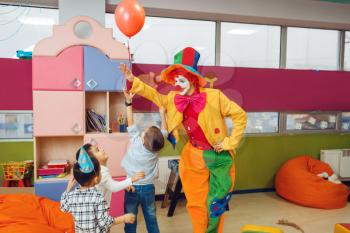Funny clown shows tricks with balloon to surprised children. Birthday party celebrating in playroom, baby holiday in playground. Childhood happiness, childish leisure
