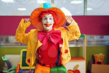 Funny clown with makeup dressed in colorful hat and costume poses in children's area. Birthday party in playroom, baby holiday in playground. Childish leisure, entertainment with animator