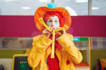 Funny clown with makeup blows into the party whistle tube. Birthday party celebrating in playroom, baby holiday in playground. Childhood happiness, childish leisure, entertainment with animator